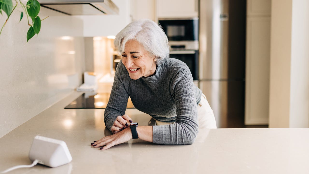 زن سالخورده در حالی که از دستگاه های هوشمند در آشپزخانه خود استفاده می کند با خوشحالی لبخند می زند.  زن مسن شاد با استفاده از دستیار خانگی برای انجام وظایف در خانه.